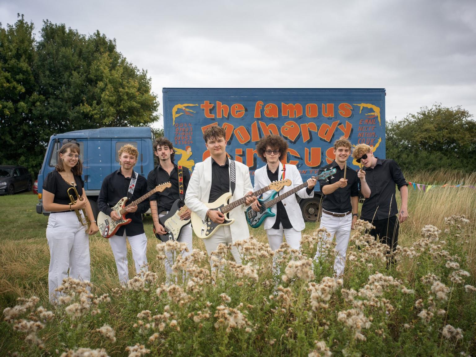 A band of young people holding electric guitars and a trumpet stand in a field of long grass in front of a bright blue and orange sign for a circus. They're wearing black shirts and white trousers.