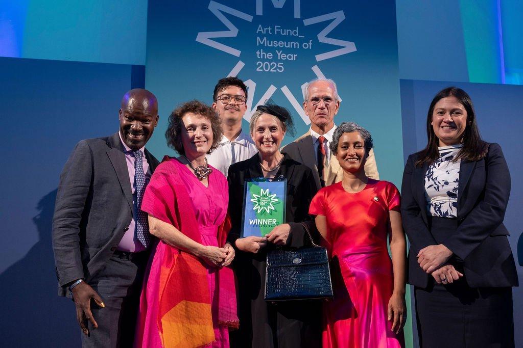 A diverse group of people on a blue-green stage hold a green "WINNER" plaque for Art Fund Museum of the Year 2025, smiling for a photo.