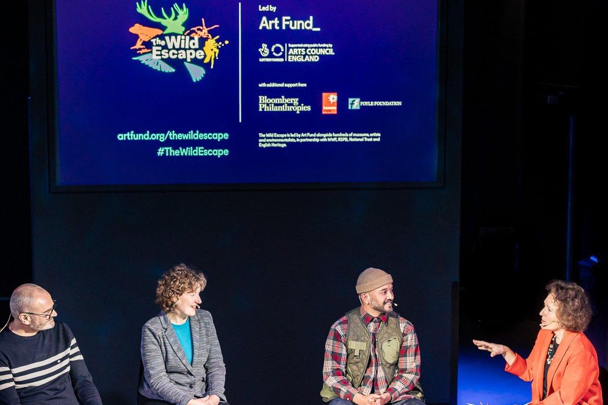A panel of four speakers sits on stage under a blue 'The Wild Escape' screen while a woman in an orange jacket gestures as she speaks.