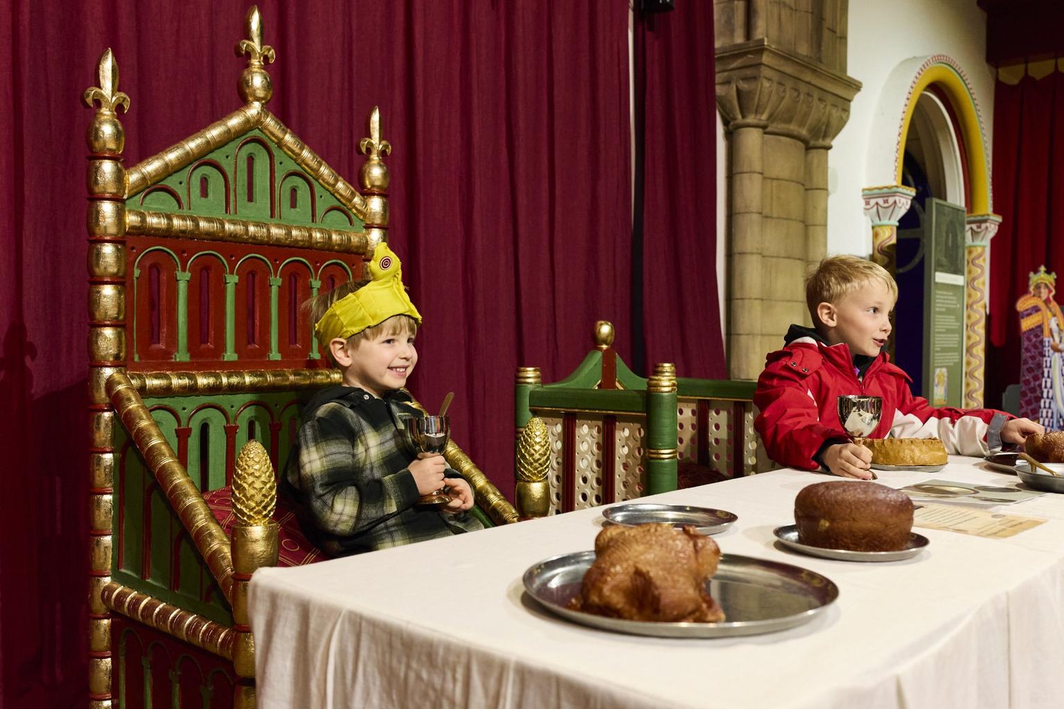 Two children sit on green and gold thrones, holding goblets at a banquet table.