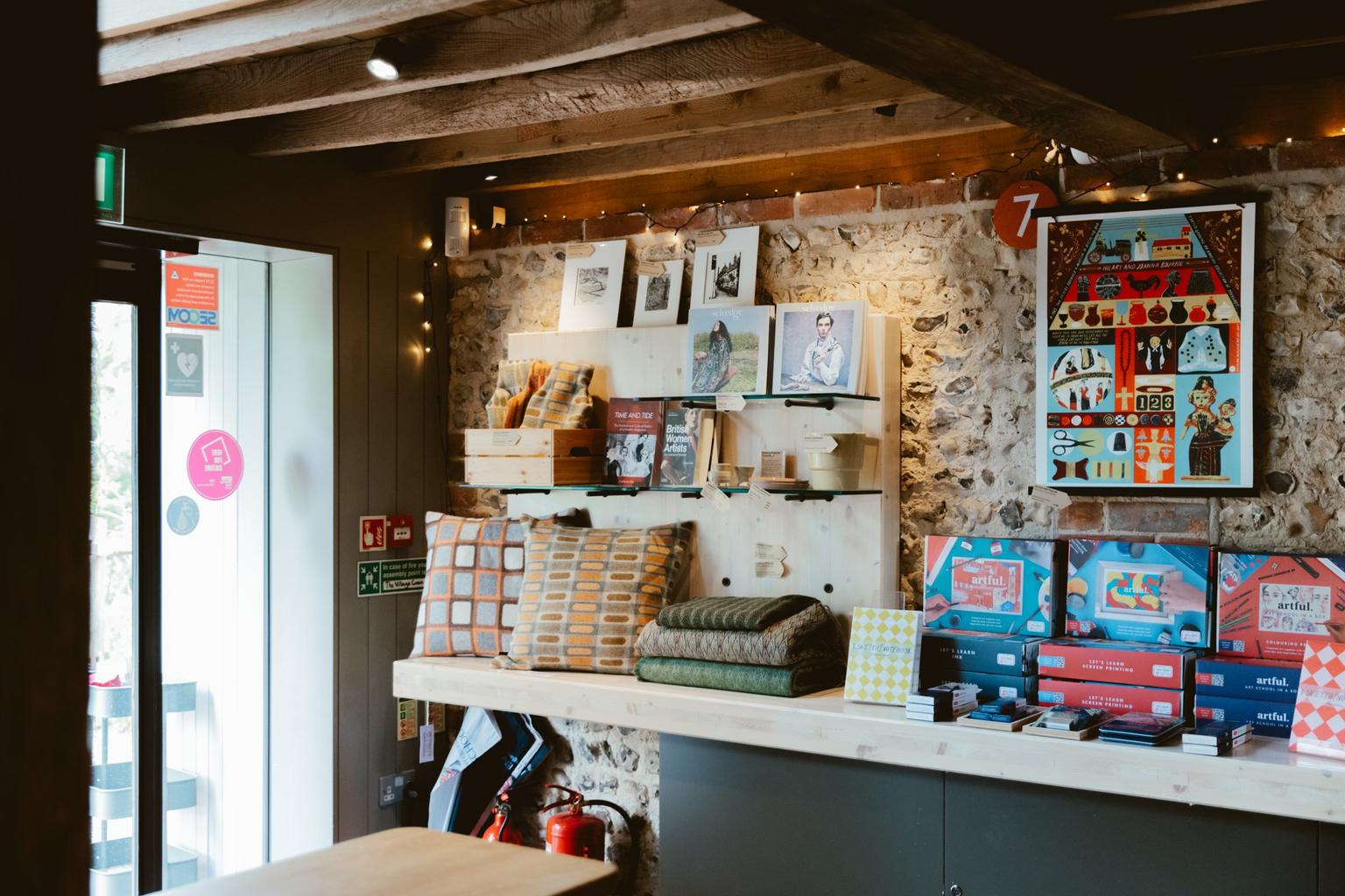 Shop counter displays patterned cushions, blankets, art prints and colorful boxed gifts against a stone wall under wooden beams decorated with festive fairy lights.