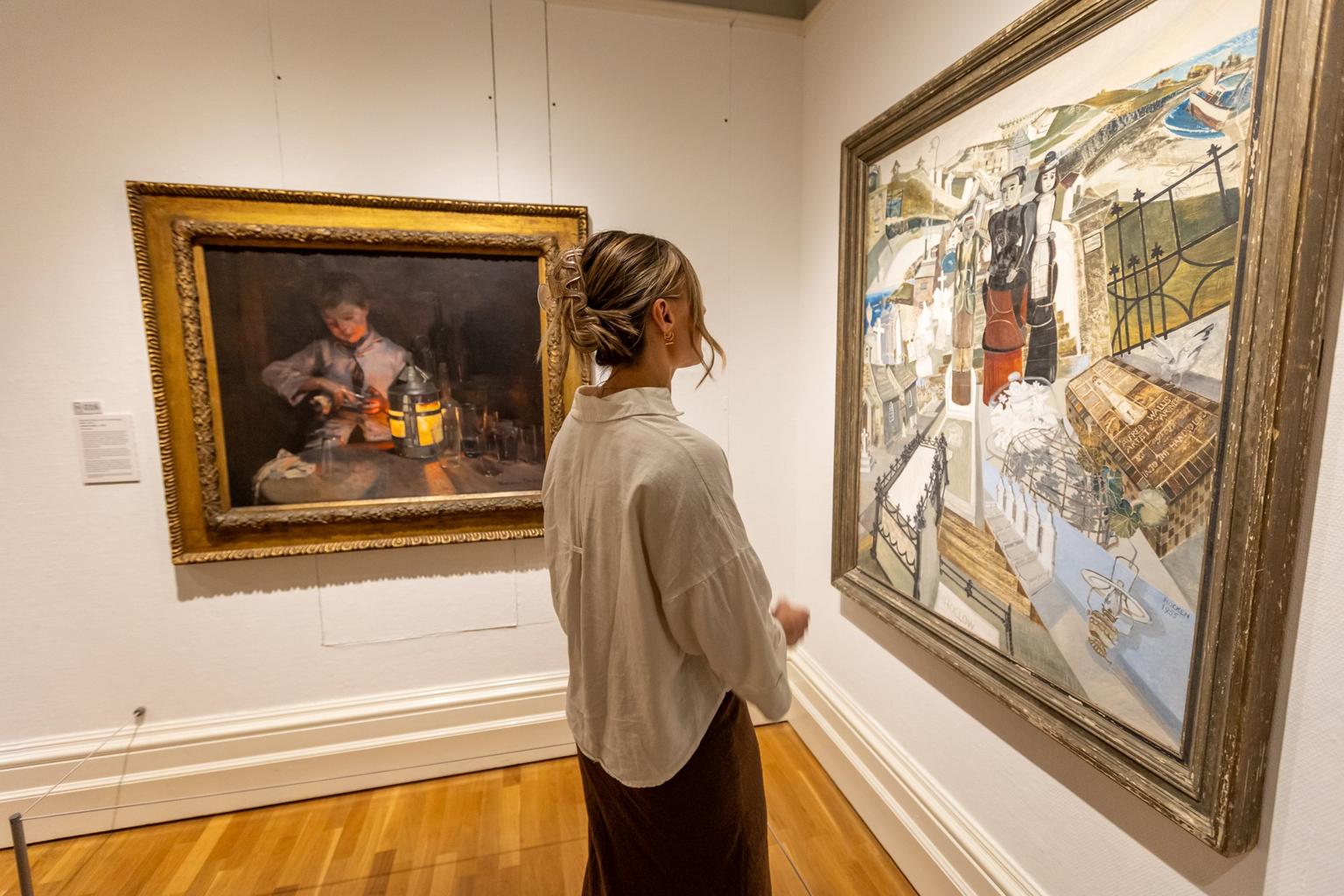 A woman in a light blouse stands in a gallery observing a large colourful painting hung beside a darker framed portrait.