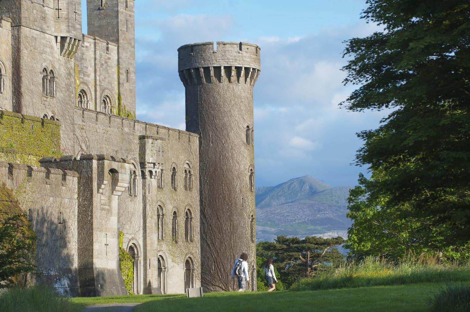 Two people walking in front of a towering castle with distant hills visible in the background