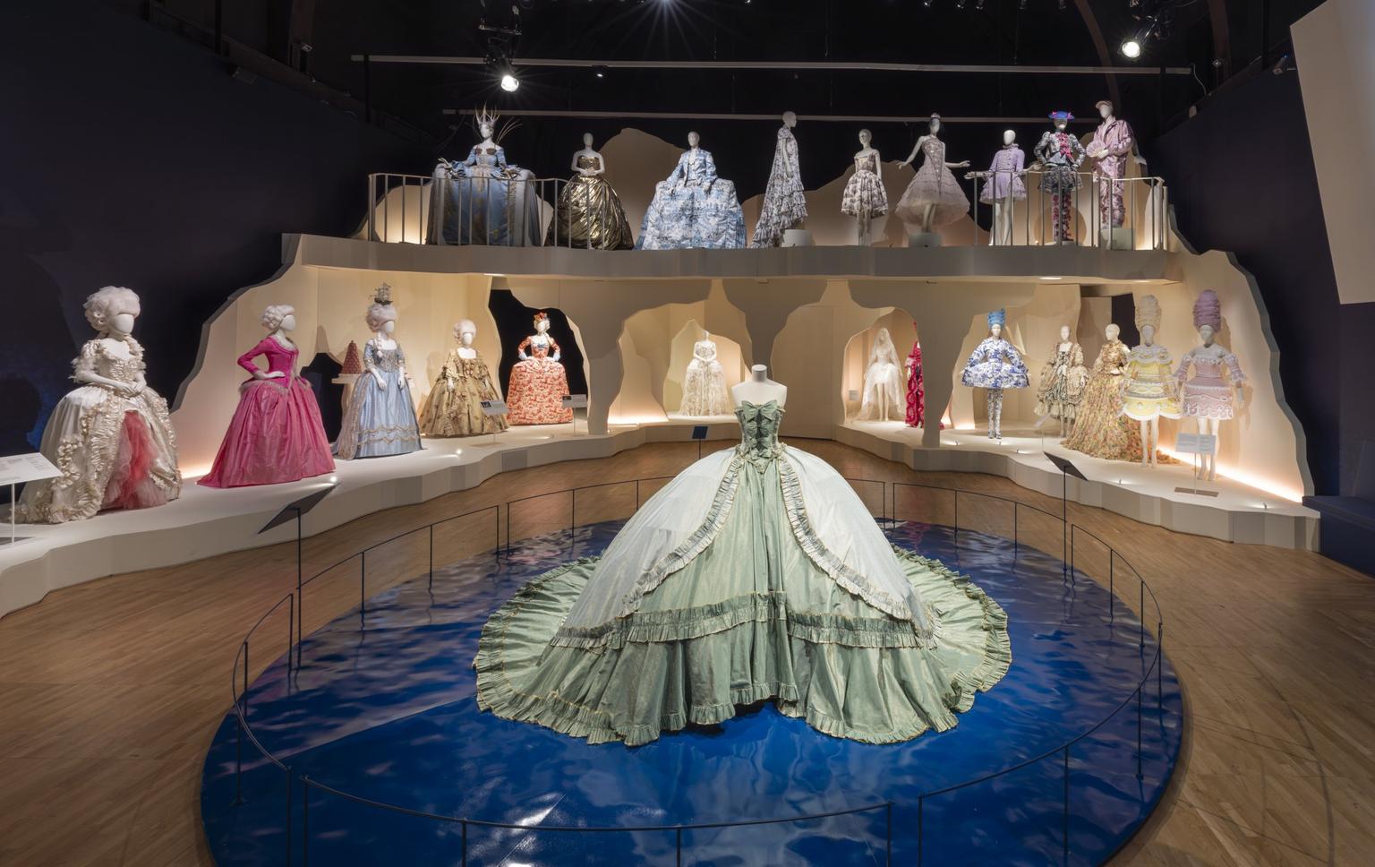 An exhibition installation space with elaborate gowns arranged in rows on two levels