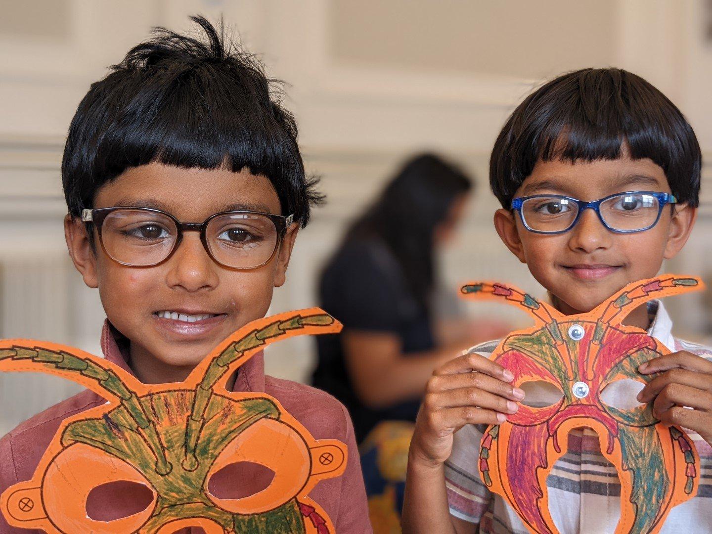 Two boys wearing glasses hold up paper insect masks that they've coloured in.