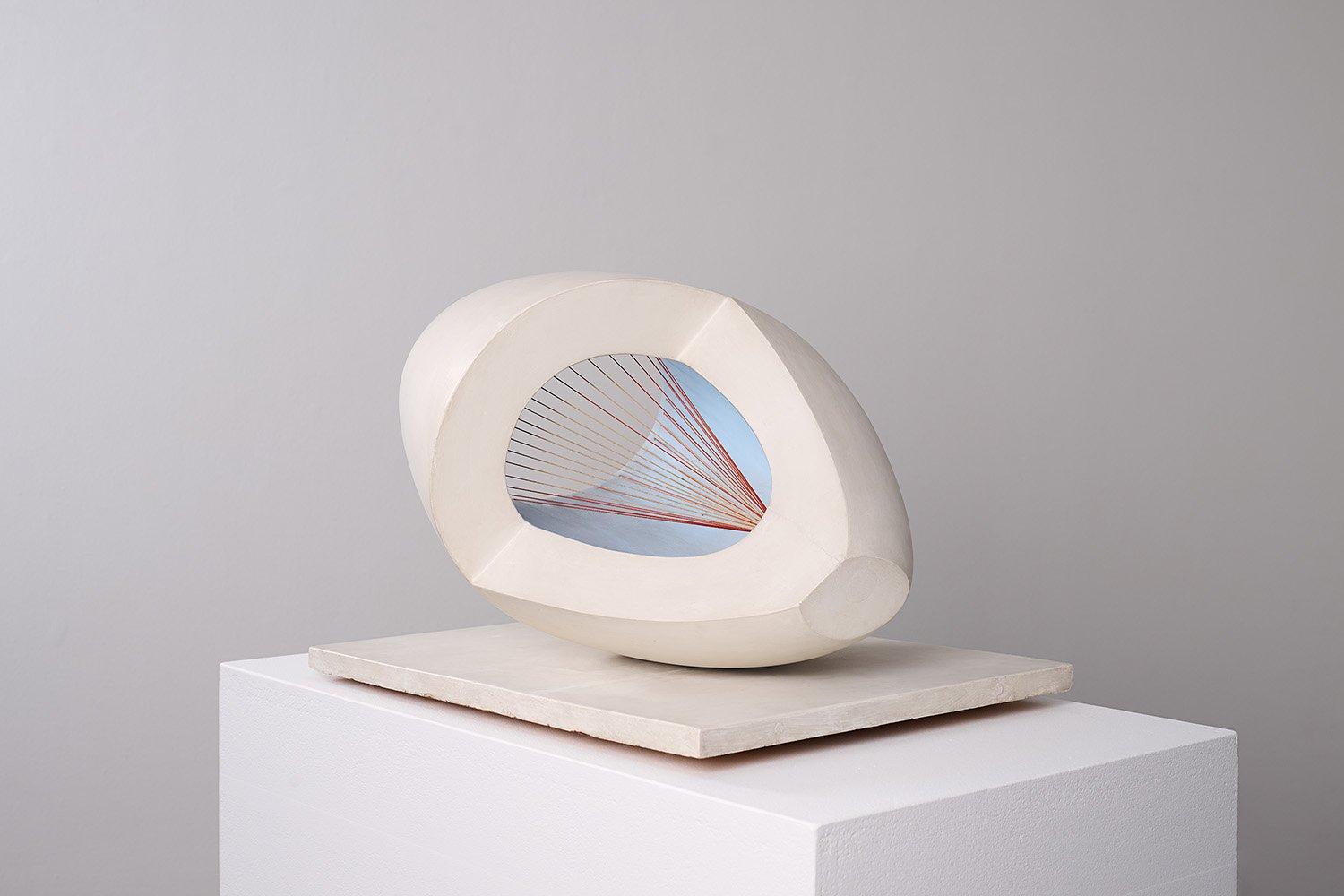 A modern abstract sculpture resembling a hollowed, egg-shaped form, partially sliced open to reveal an interior painted sky blue. Inside, a series of taut red strings radiate from a single point at the base toward the open edge, creating a geometric fan-like pattern. The sculpture rests on a white plinth against a neutral grey background.