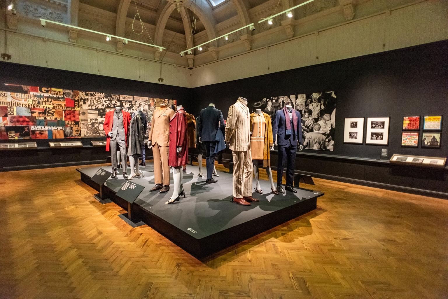 In the centre of the gallery space is a black raised platform with mannequins wearing an array of 60s suits, dresses and coats; on the walls hang black and white photographs and colourful posters from the time.