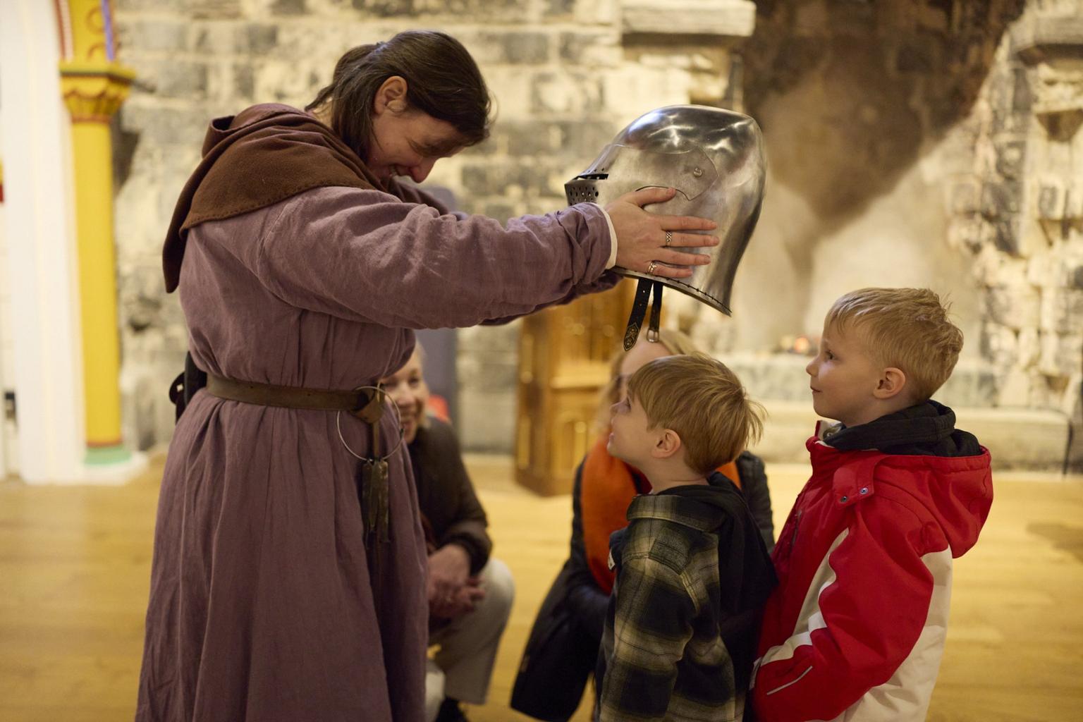 A person in a purple medieval robe holds a shiny metal helmet over a young child's head inside a castle.