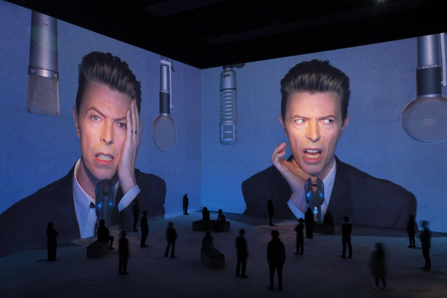 Gigantic projections of David Bowie's expressive face singing into microphones dominate a dark exhibition space with silhouetted visitors.