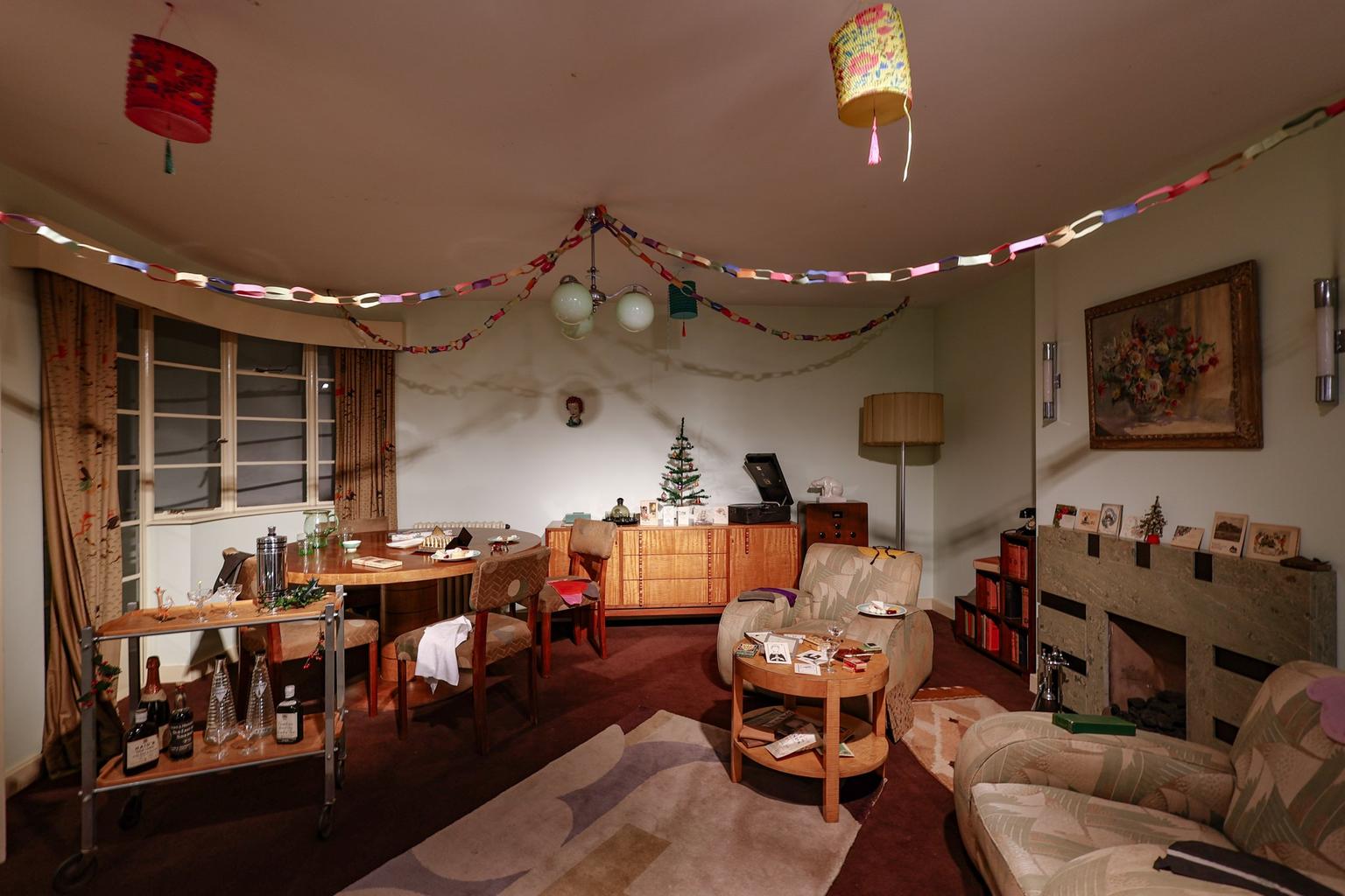 A mid-century living room decorated with colorful paper chains and a small Christmas tree, featuring wood furniture, beige armchairs and a drinks trolley.