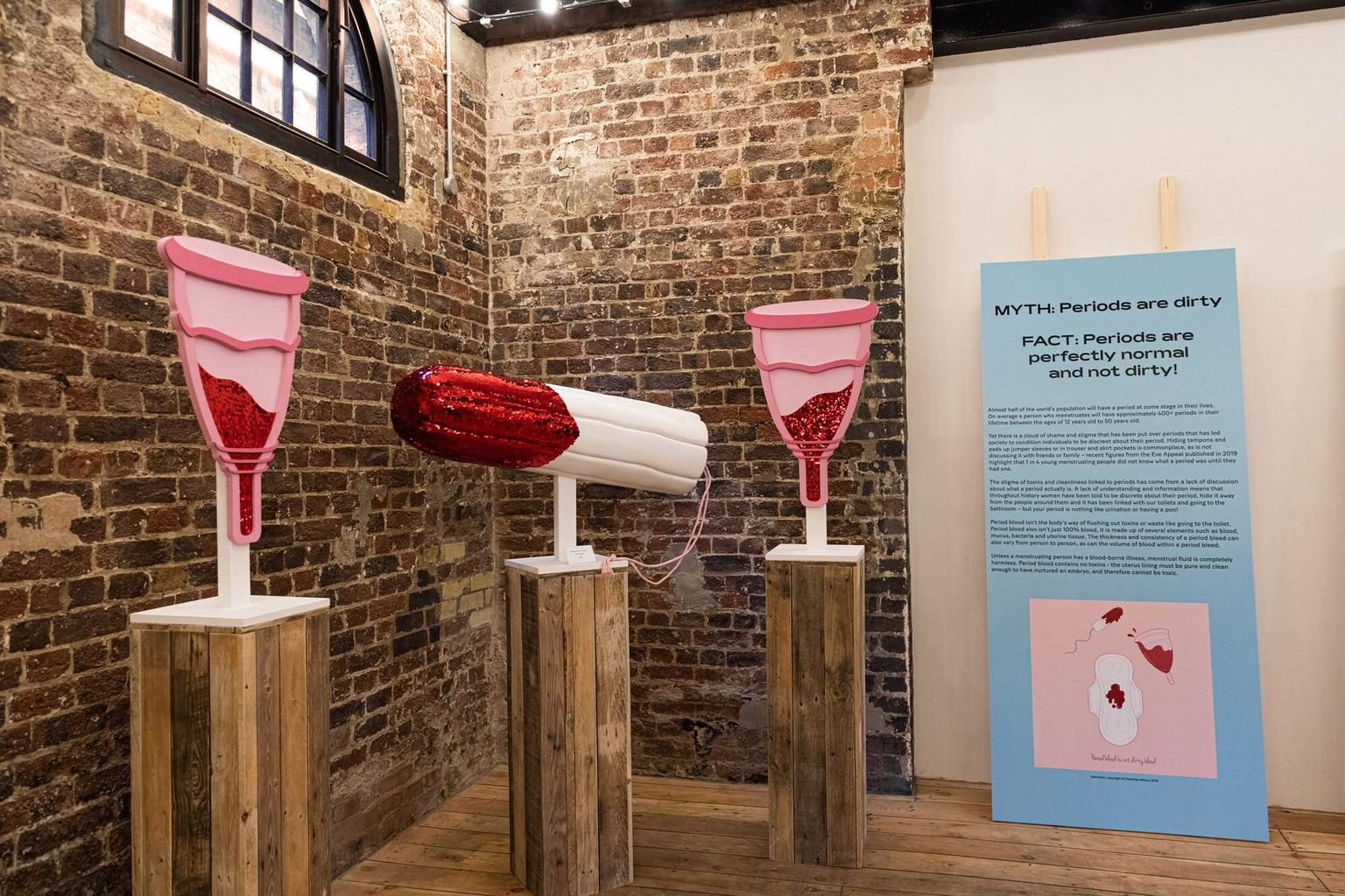Three plinths holding sculptures of a tampon and two mooncups featuring blood in red glitter
