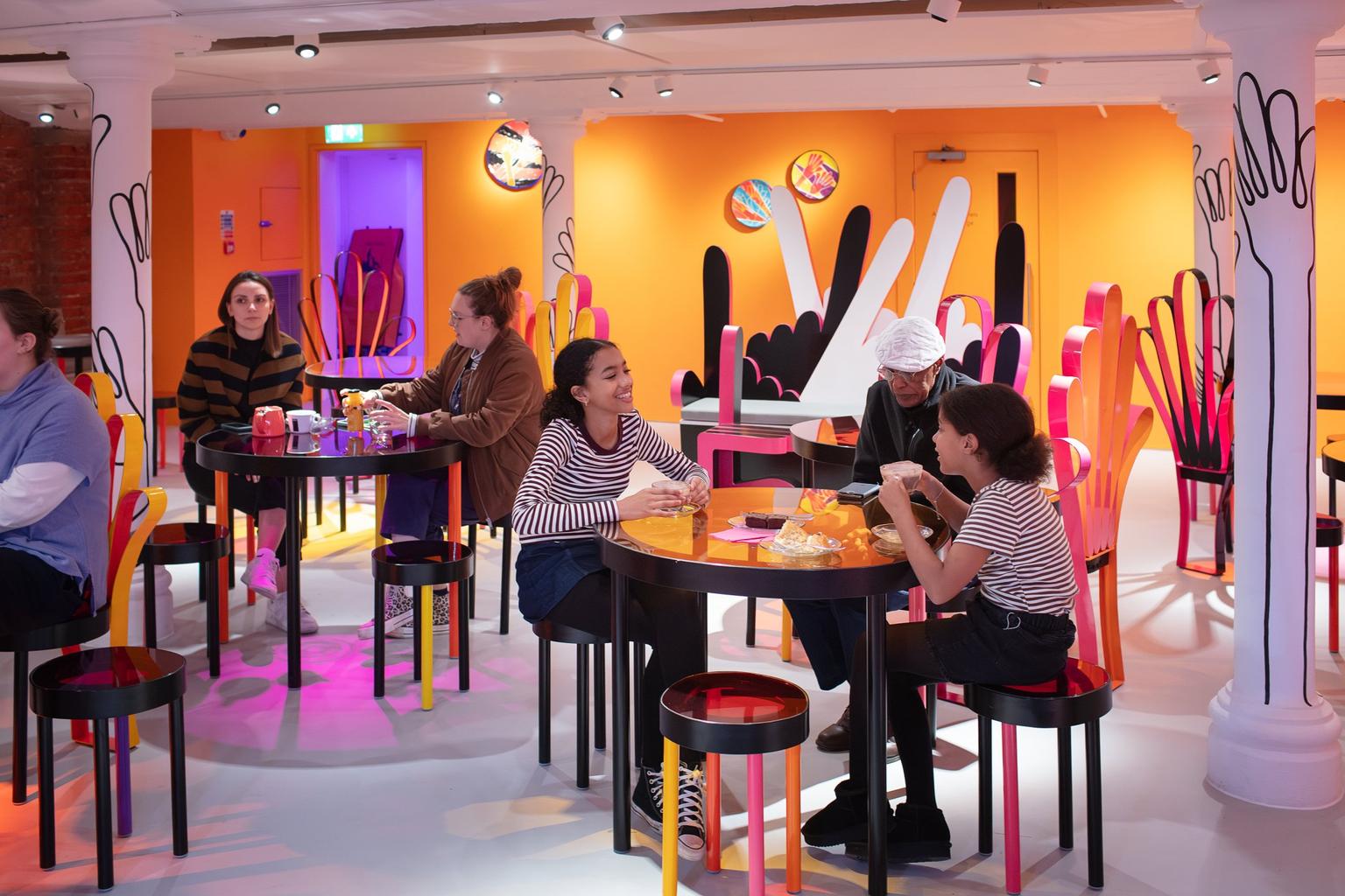 Visitors sitting and eating in a vibrant café with orange walls, tables and chairs with orange, pink and yellow legs.