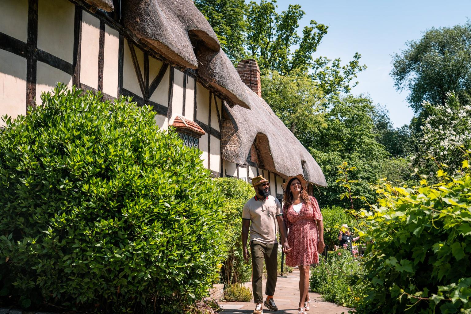 A man and woman walking along a path in front of a thatched cottage surrounded by greenery
