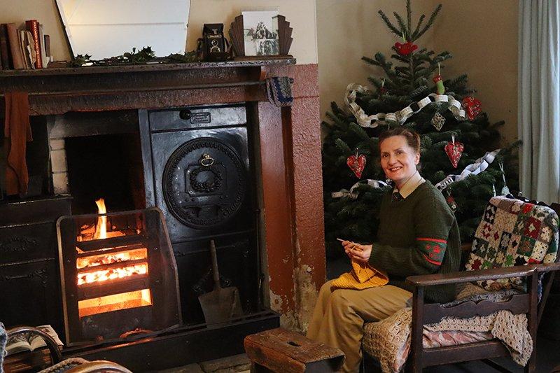 A woman in a green jumper knits yellow yarn while seated by a roaring fireplace beside a decorated Christmas tree with red heart ornaments and a paper garland.