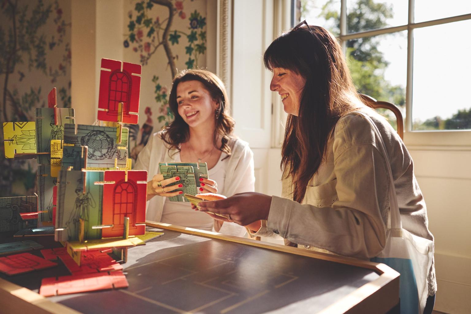 Two women sit by a sunlit window smiling as they build a colourful red and green 3D card structure while holding game cards.