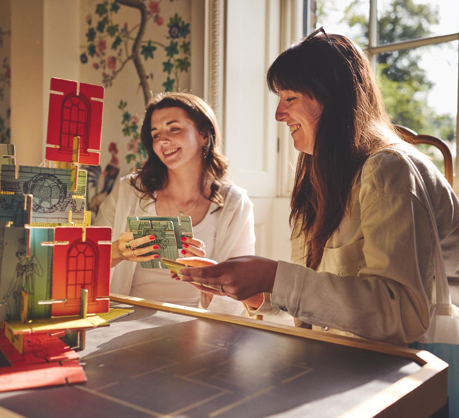 Two smiling women play a colorful modular tabletop building game at a sunlit table by a window.