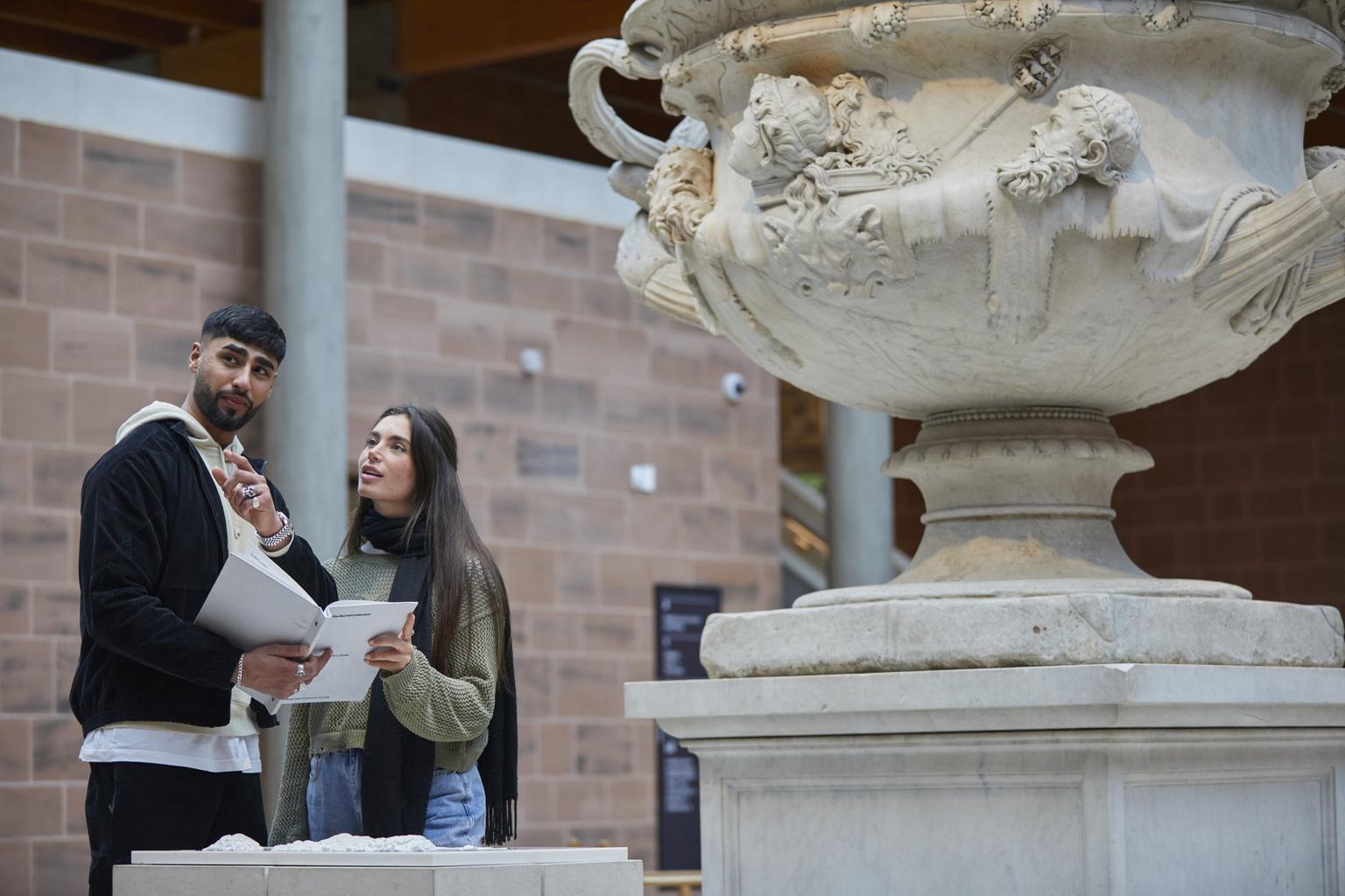 Two visitors stand in a courtyard near the marble Warwick Vase