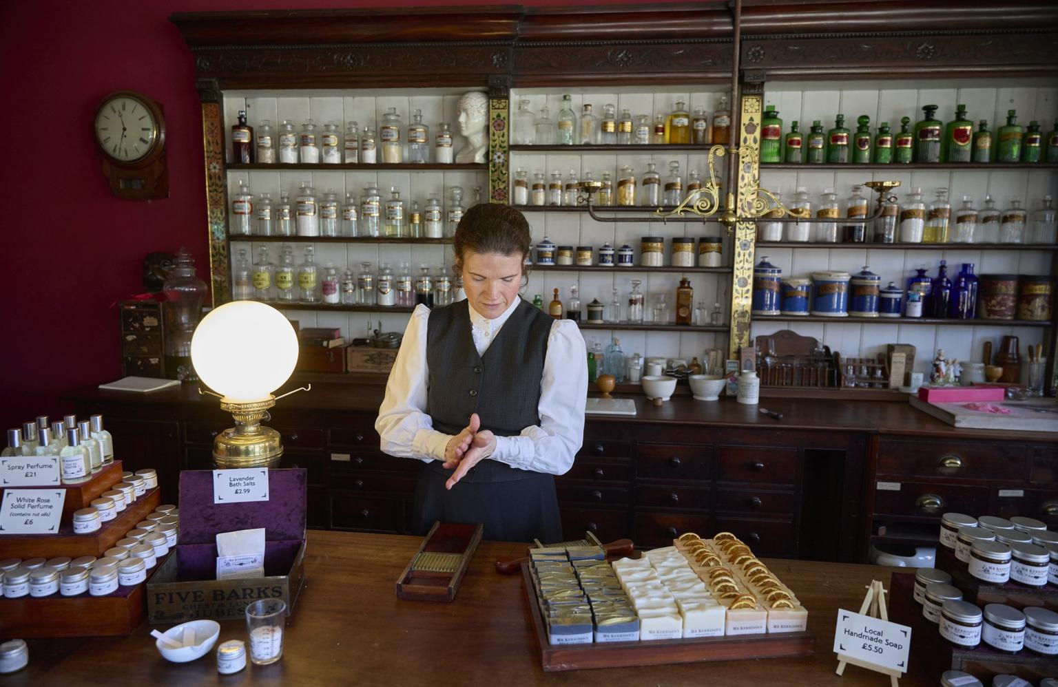 A woman in an Edwardian costume sells soaps, perfumes and bath salts in a traditional chemist. Behind her are shelves of glass bottles holding a range of lotions and ointments.