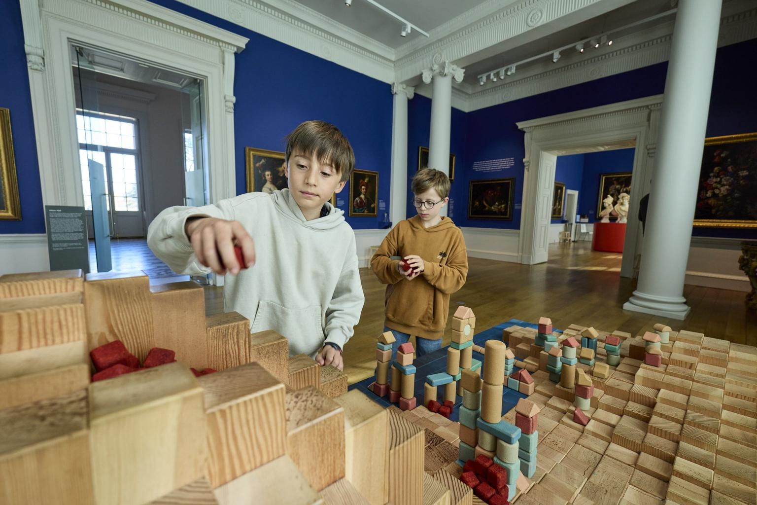 Two boys playing with wooden building blocks in a blue walled gallery space with Old Master paintings hanging on the walls.