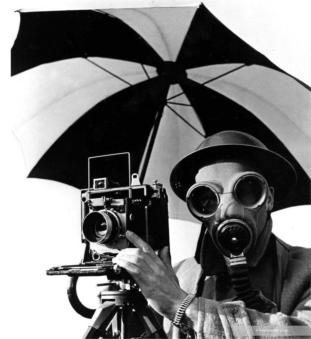 Black-and-white photo of a photographer wearing a gas mask and round goggles operating a vintage camera under a large umbrella.