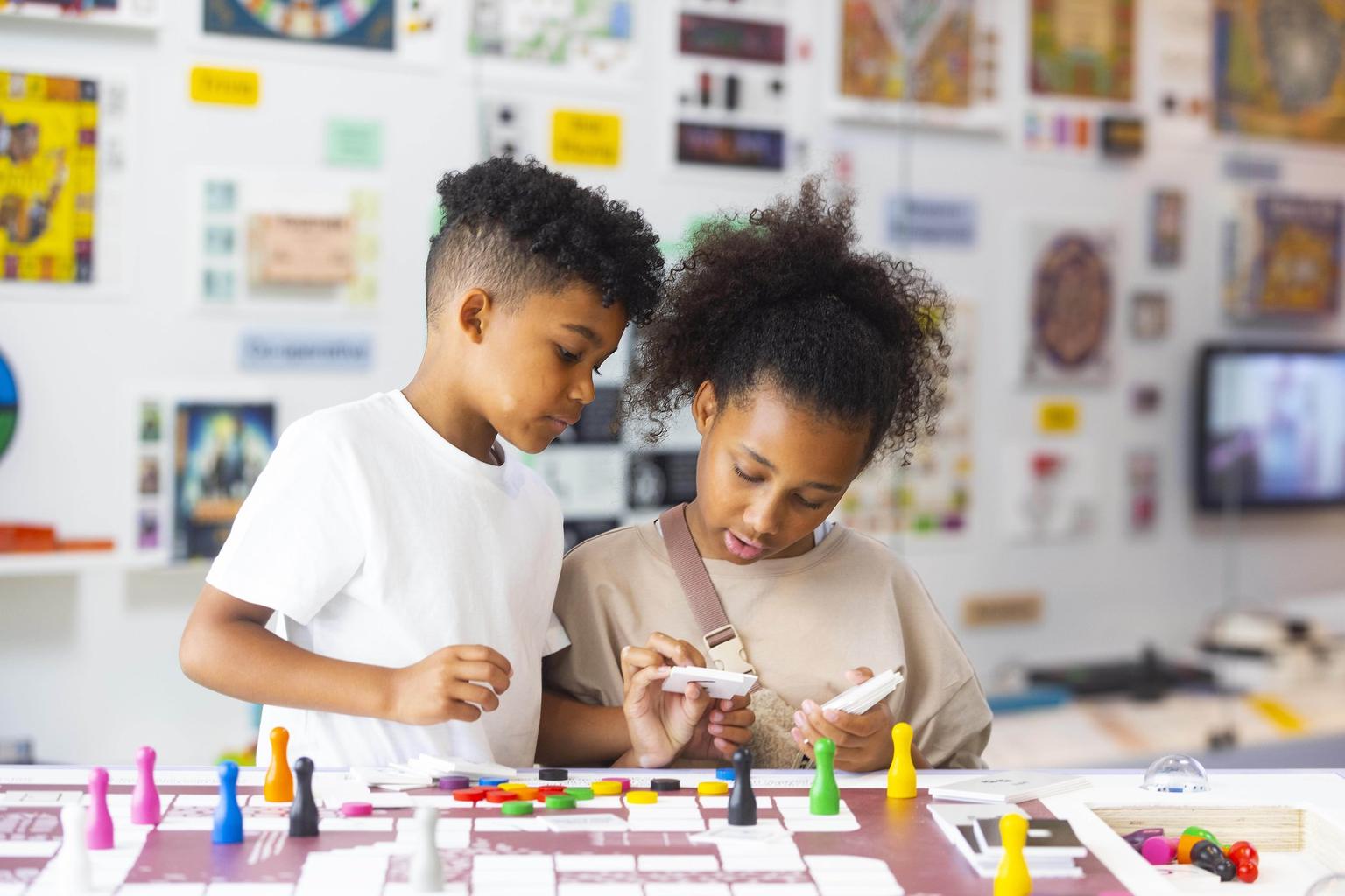 Two children examine cards while playing a colorful board game on a white table in a bright classroom.