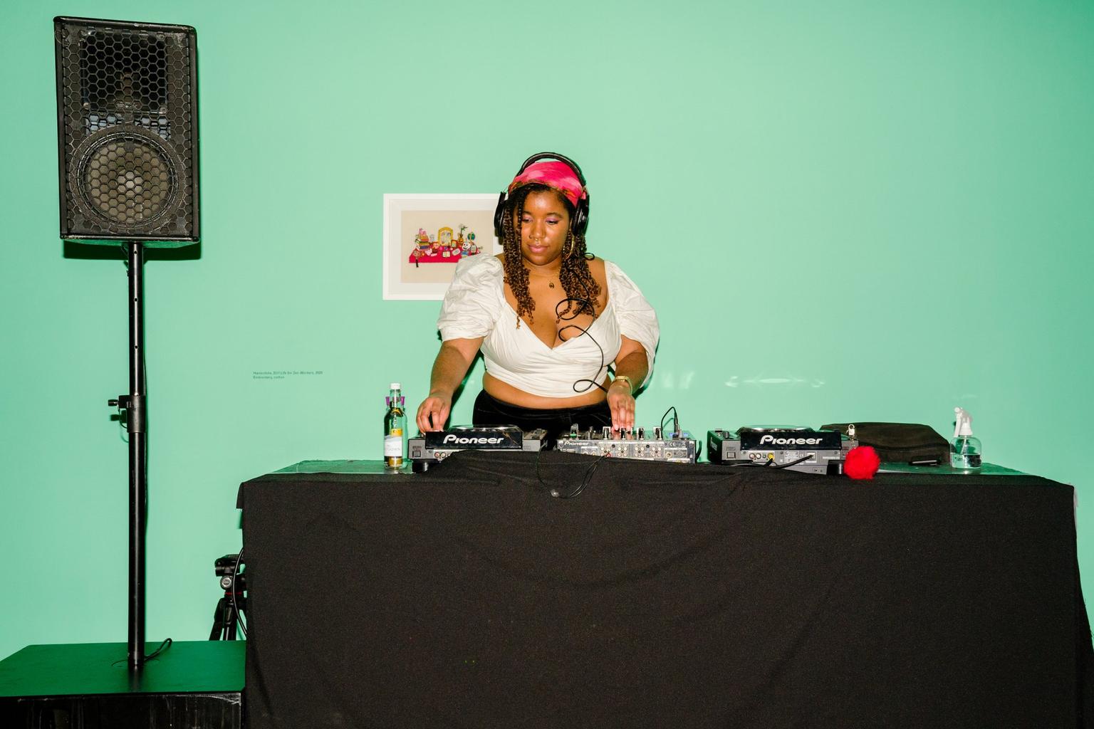 A DJ in a white top and pink headband mixes music on DJ decks in front of a mint green wall.