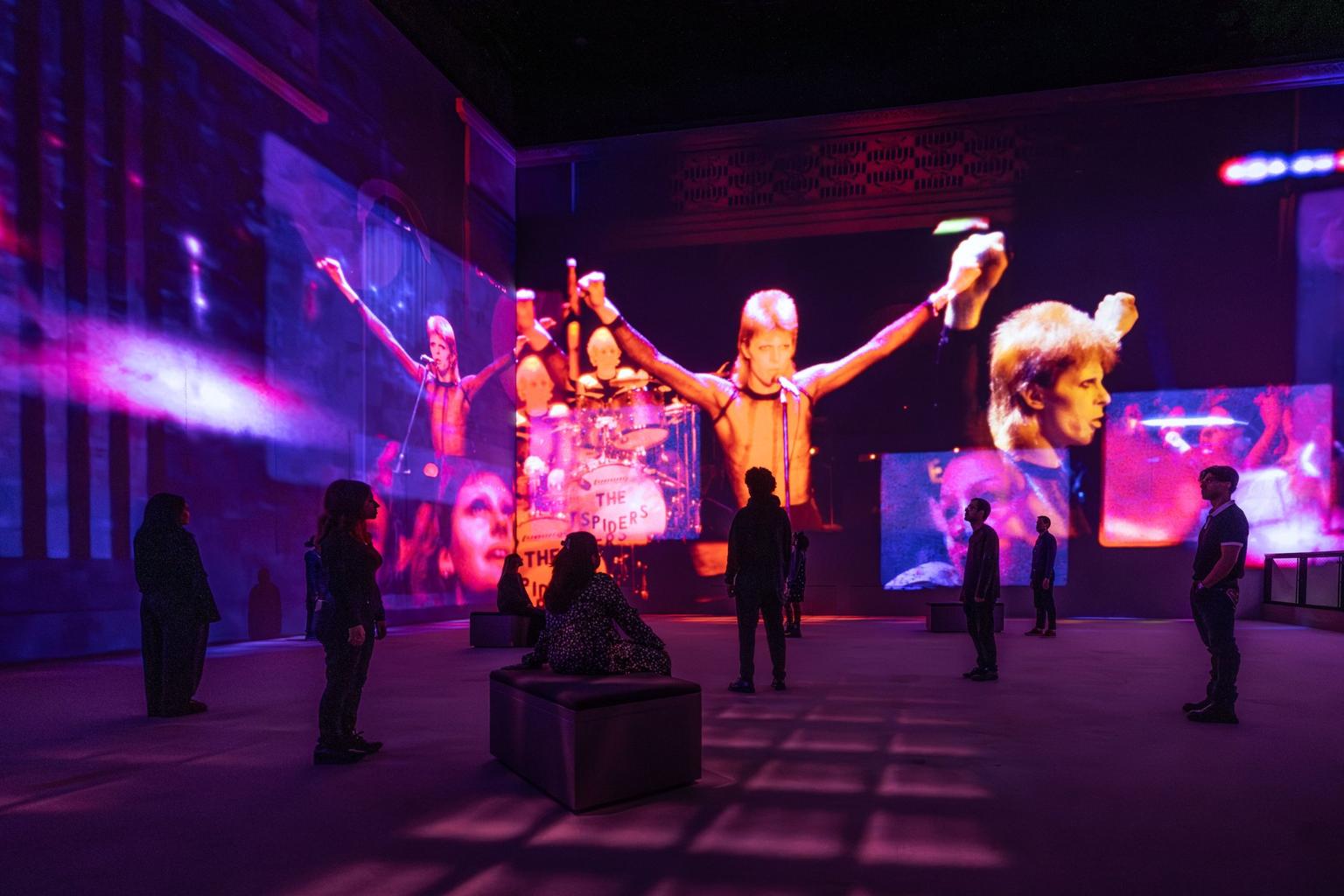 Visitors stand in a gallery watching purple, pink and orange projections of David Bowie performing across the walls.