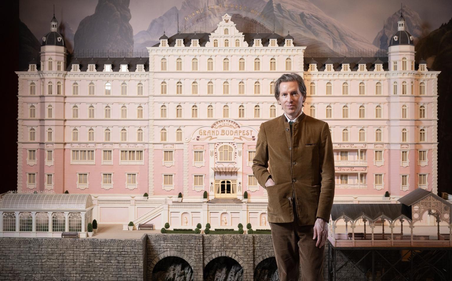 Wes Anderson stands in front of a large pink model of the Grand Budapest Hotel.