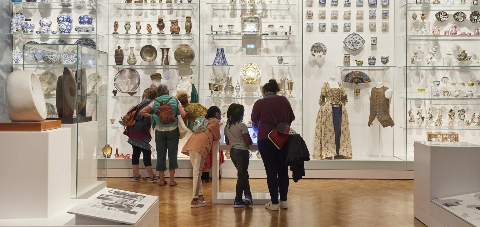 Two groups of people stand in front of a wall of different artefacts