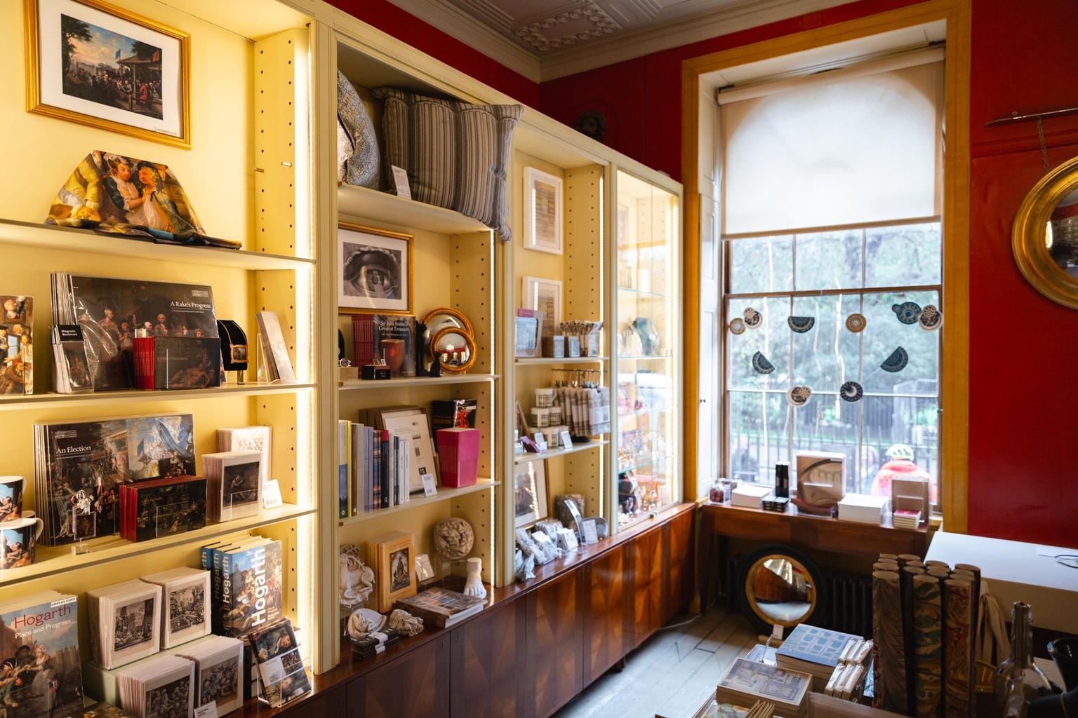 A cozy museum gift shop with illuminated shelves displaying books, souvenirs, and decorative items against deep red walls and a window decorated with paper garlands.