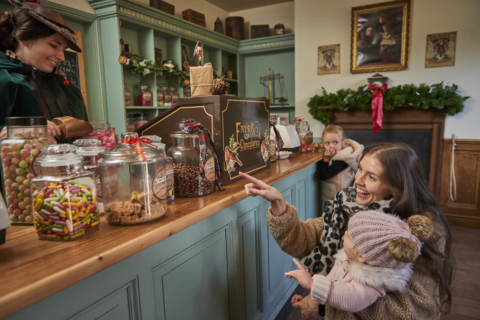 Woman in a fur coat points at jars of sweets on a teal Victorian shop counter while holding a toddler in a pink hat as a shop assistant measures sweets.