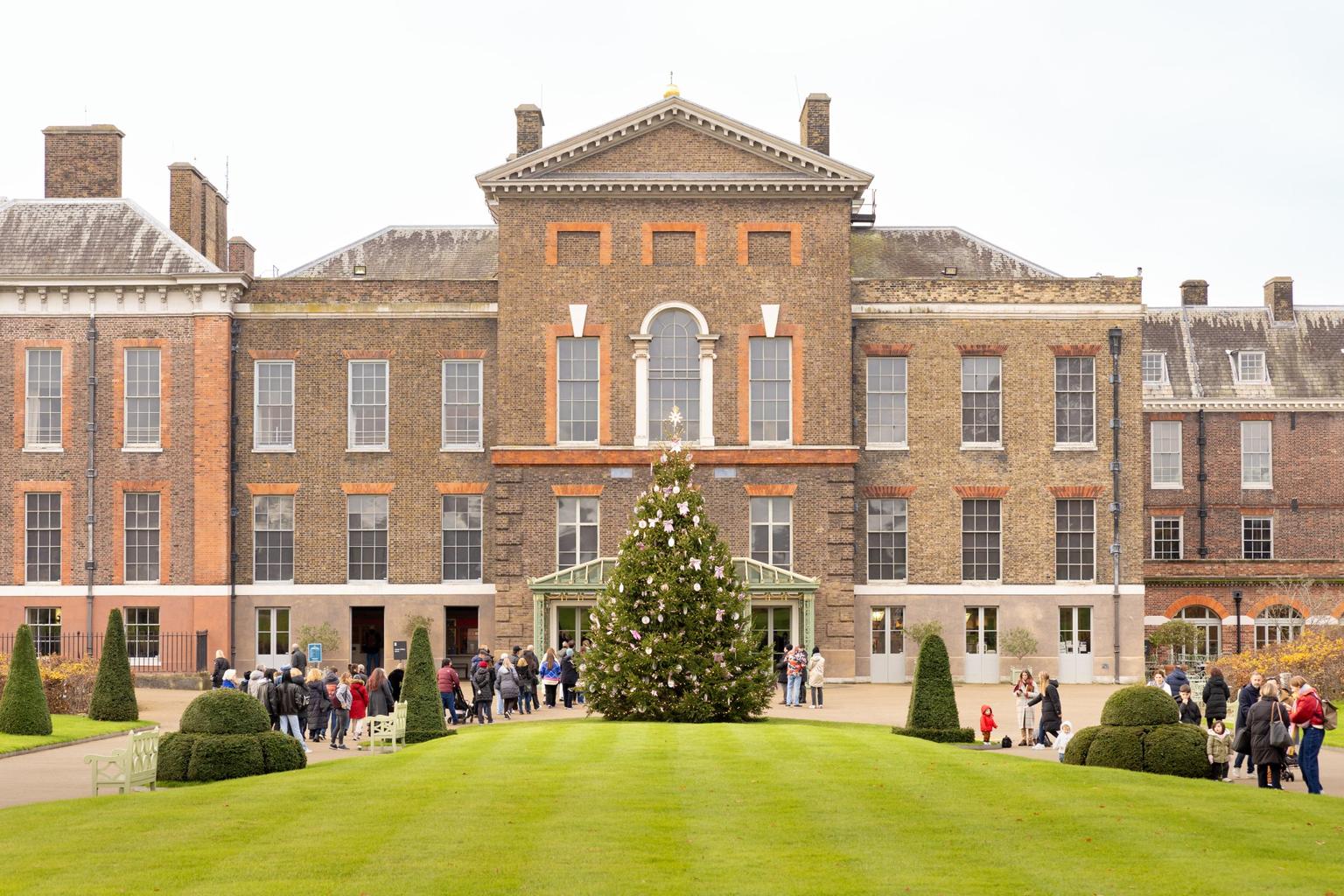 A large decorated Christmas tree stands on a green lawn in front of a historic palace as visitors gather nearby.