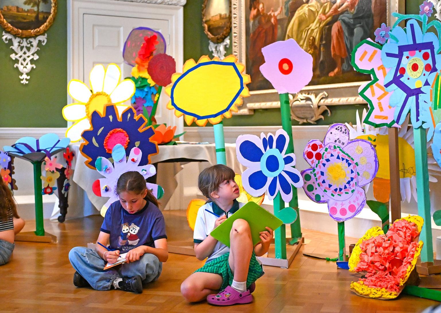 Two children sit on a wooden floor sketching amid oversized colourful model flowers in a gallery room.