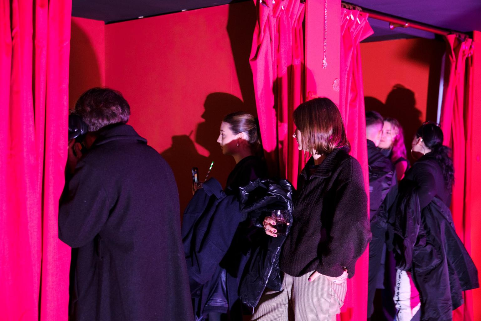 A group of people in dark coats are watching something out of shot, beside bright pink curtains and red walls.