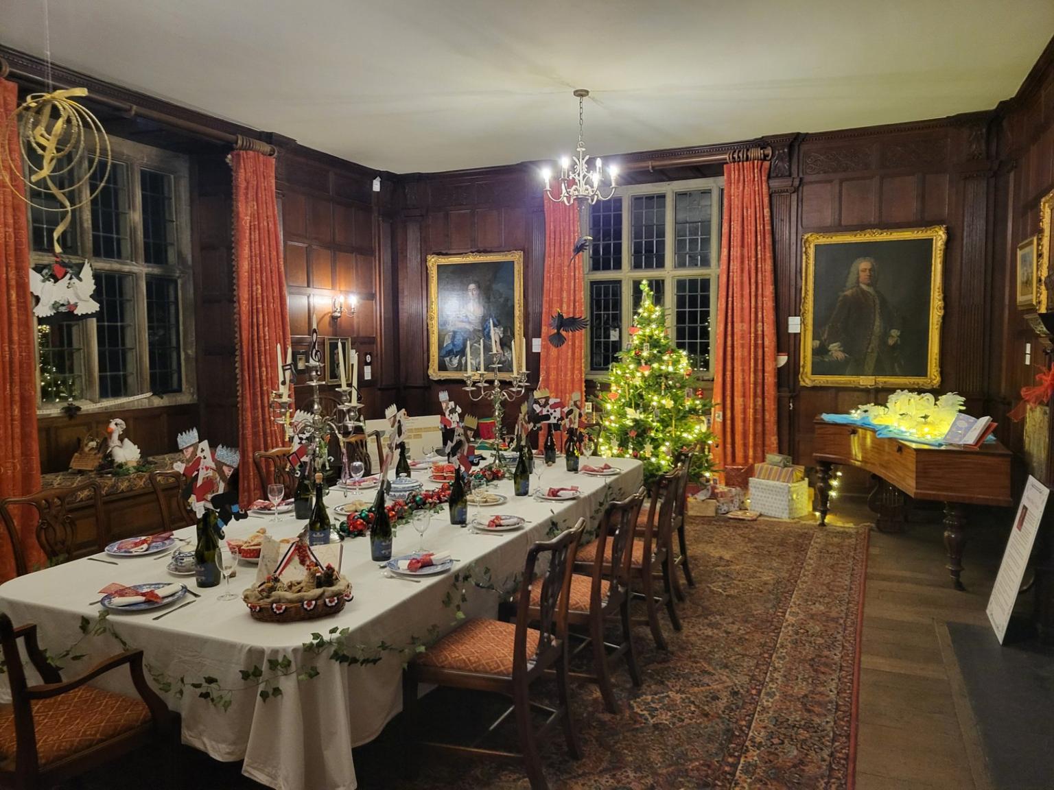 A long dining table set for Christmas with a white tablecloth, festive place settings and a lit Christmas tree in a wood-paneled room with red curtains.