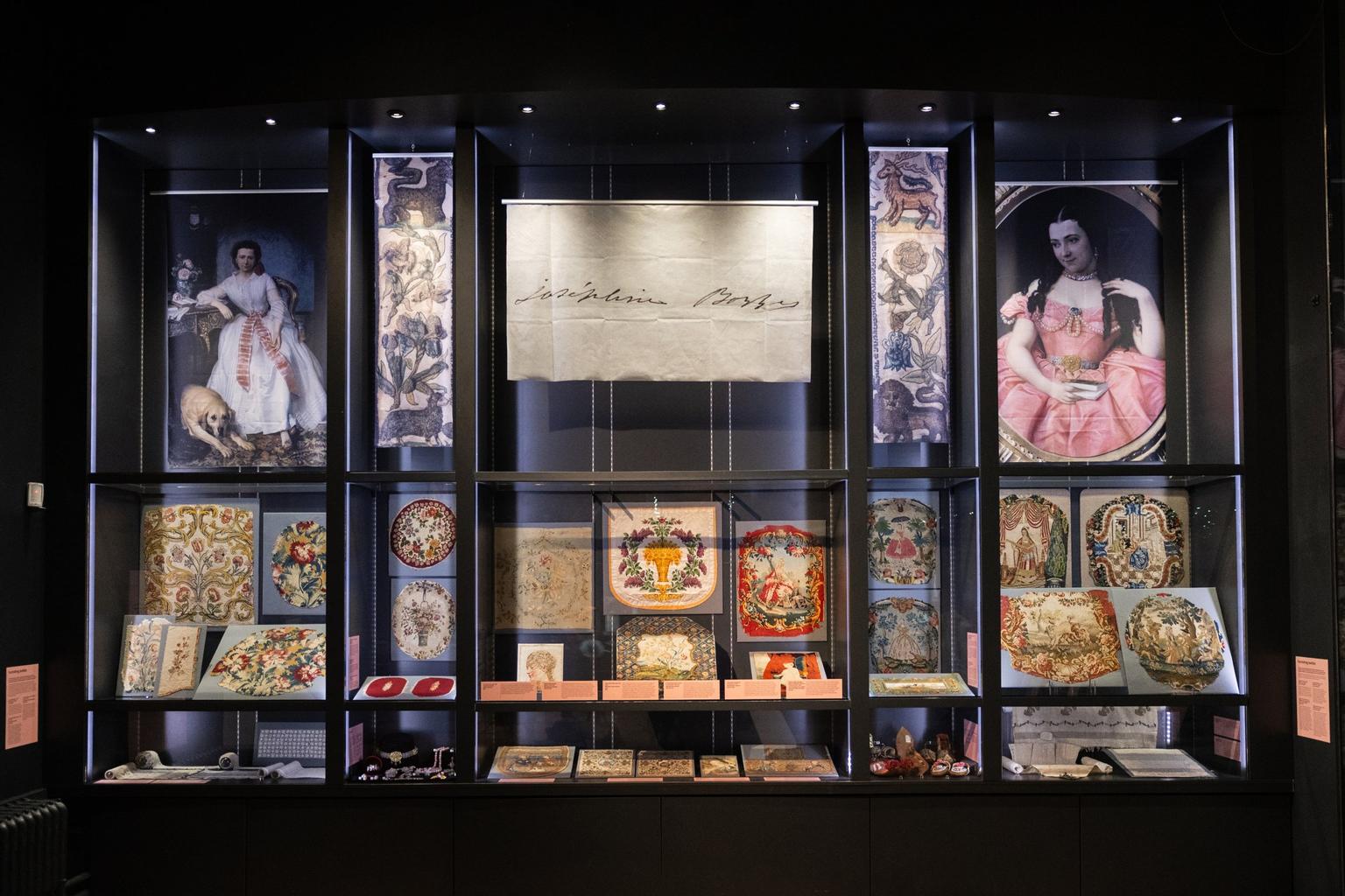 A museum case filled with hanging fabrics and two portraits of a woman with brown hair
