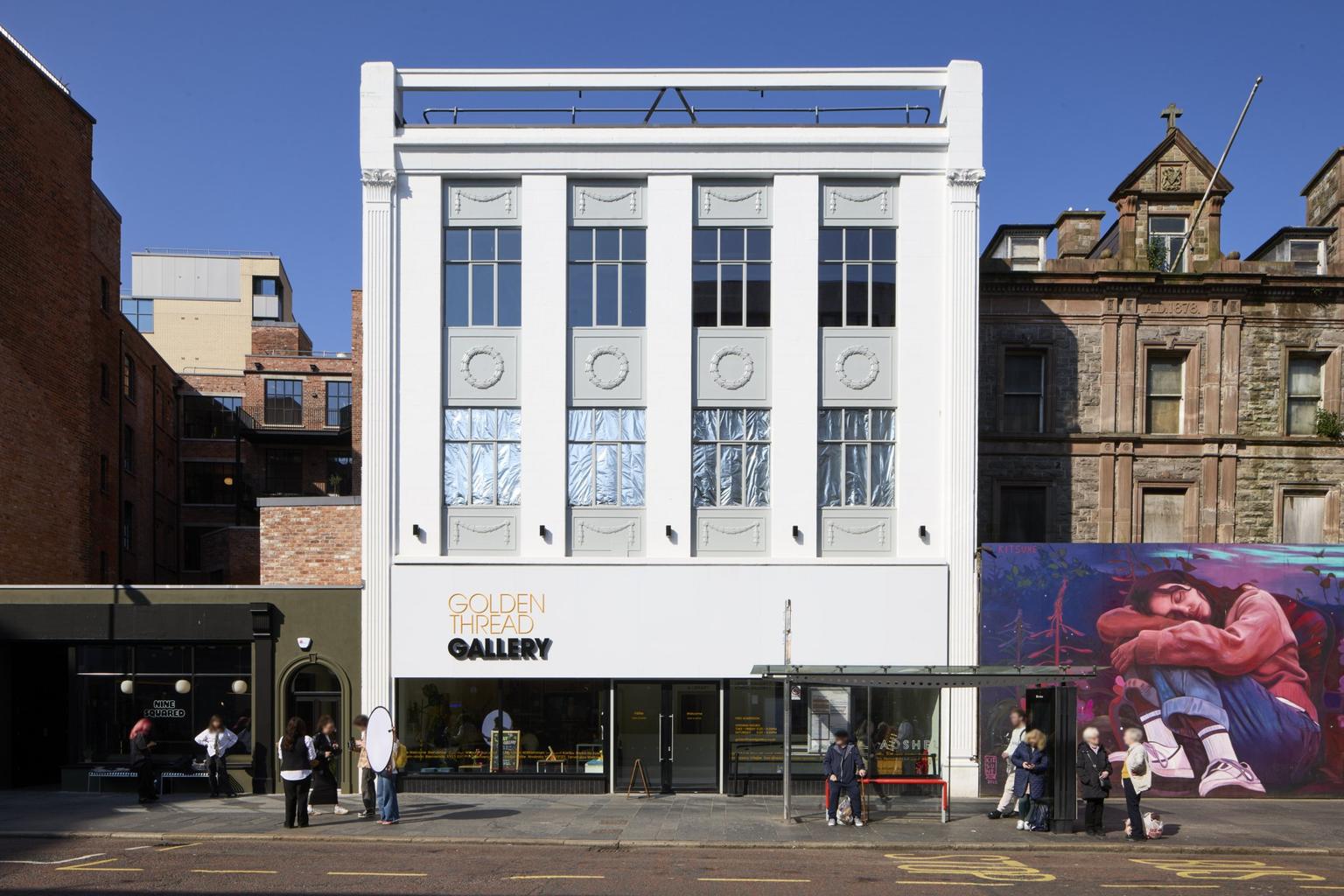 A tall white gallery building with a horizontal glass front across the bottom floor and a sign reading 'Golden Thread Gallery' in yellow and black.