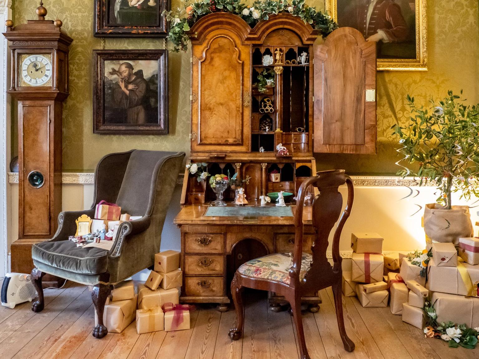 An antique wooden secretary desk with its top cabinet open sits beside a green upholstered armchair and piles of wrapped gifts in a historic parlour.