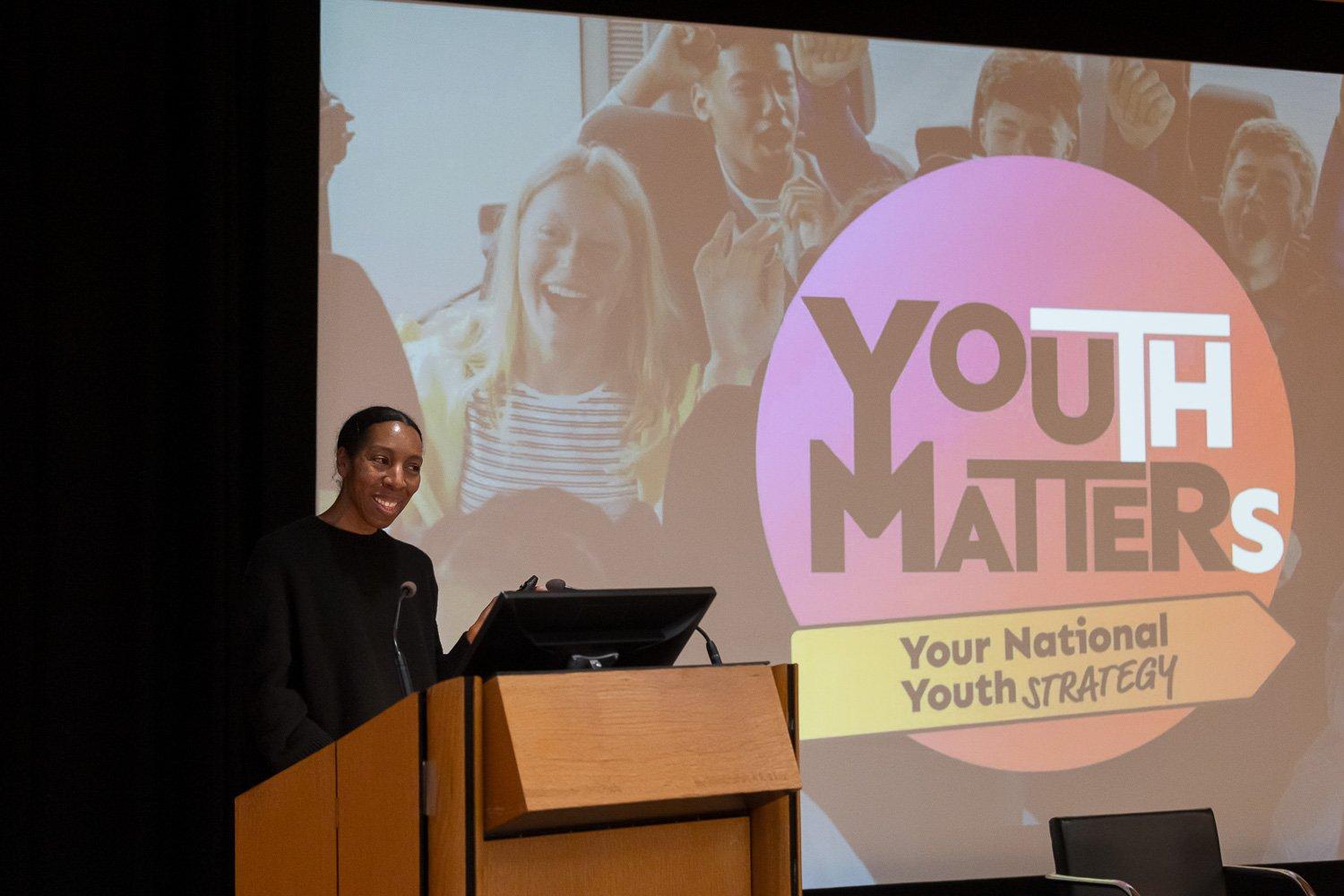 A woman speaks at a wooden podium beside a large pink and orange 'Youth Matters' logo projected on a screen.