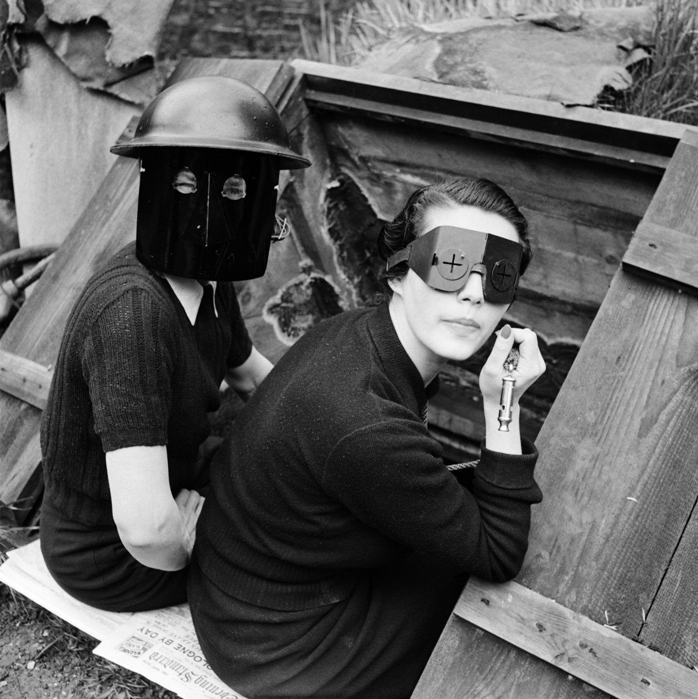 A black and white photograph of two women sitting in a wooden trench and wearing protective headgear, one with a full black mask and the other wearing crossed-eye goggles while holding a whistle.