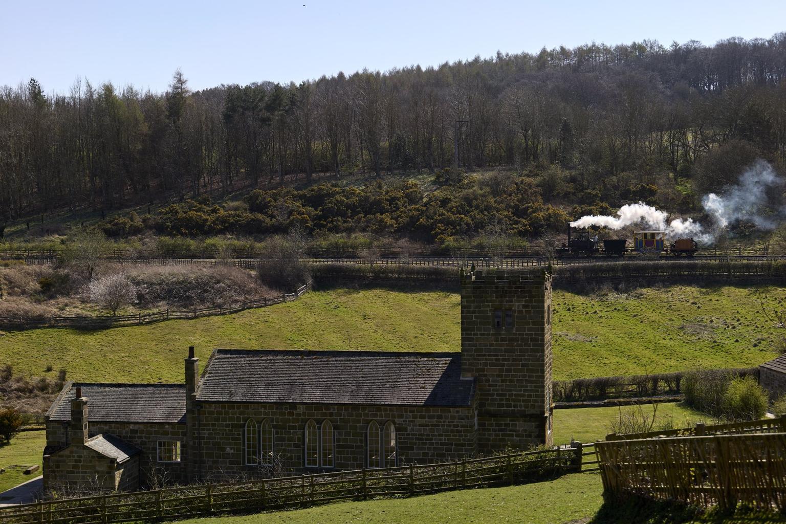 A steam train travels through the countryside with trees on its right, and green fields with a church building to its left.
