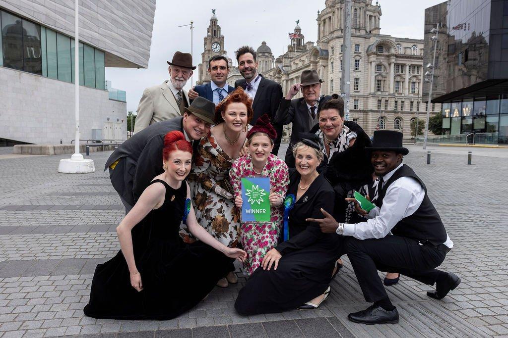 A group of people pose outdoors, centre person holding a "WINNER" sign, with city buildings behind them.