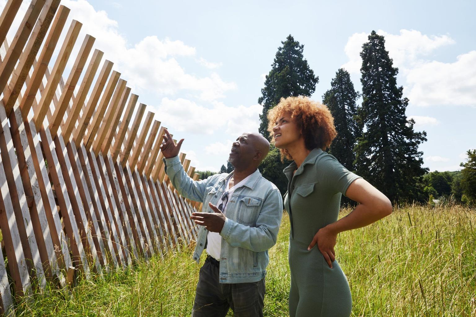 Two people stand among long grass, looking up at a sculpture of intersecting wooden beams.