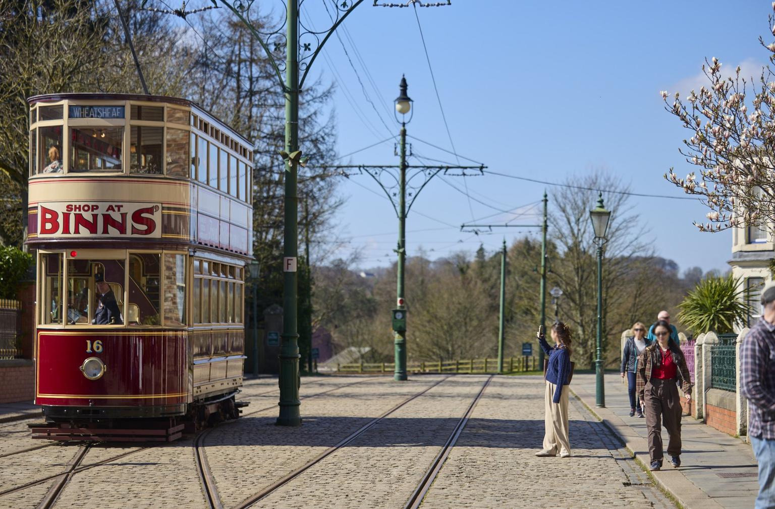 A traditional red and cream tram travels through cobbled streets with old green street lamps.