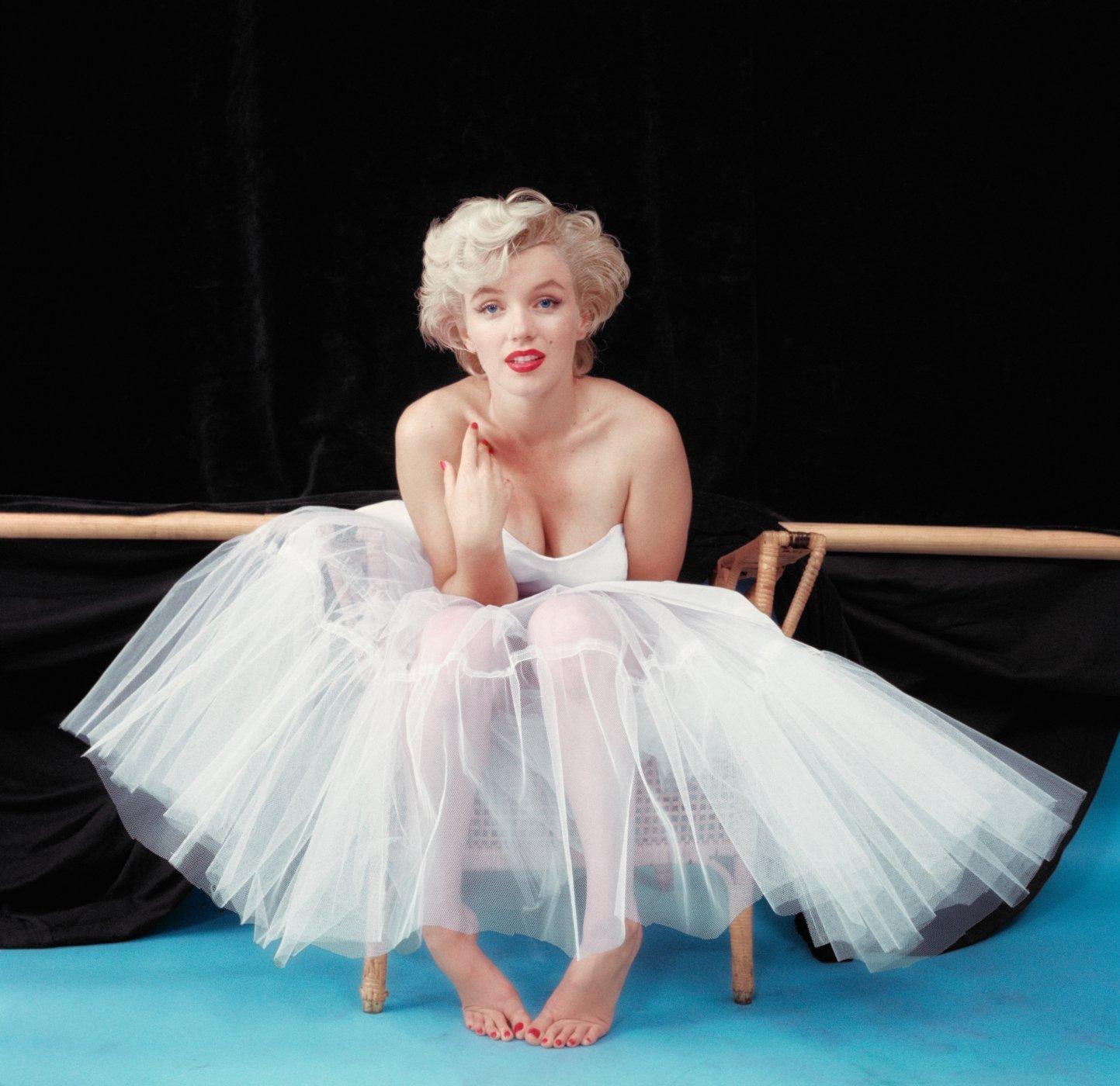 Marilyn Monroe in a white ballerina dress sits barefoot on a wicker chair against a dark backdrop and sky blue floor, looking toward the camera. Her lips and nails are painted red.