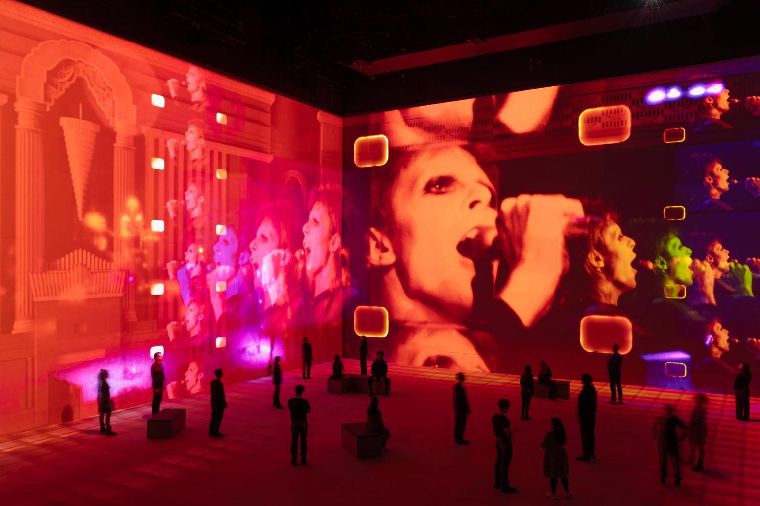 Visitors watch an immersive projection tinged with red and orange of David Bowie singing into a microphone across the gallery walls.