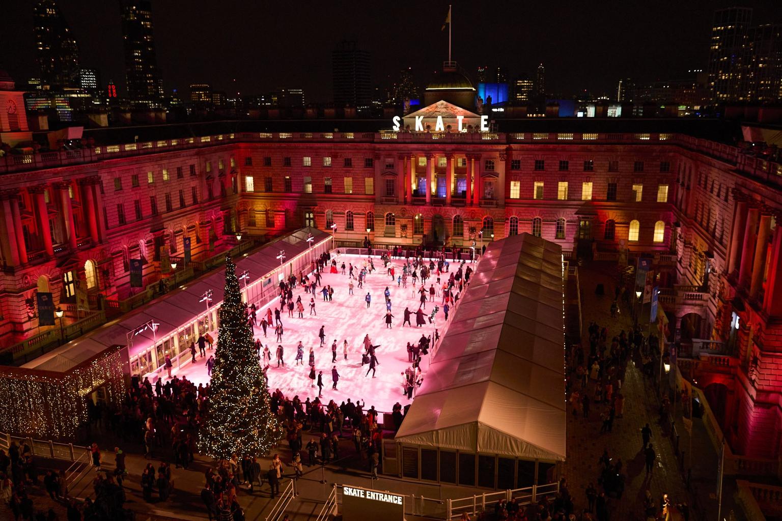 Aerial view of people ice skating on an outdoor ice rink in a grand, illuminated courtyard beside a decorated Christmas tree at night.