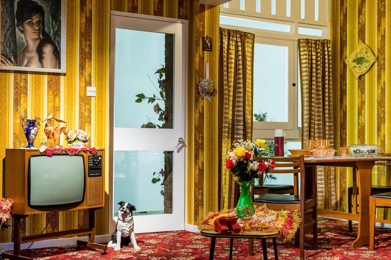 A 1970s front room with yellow striped wallpaper, a red floral carpet and 1970s furniture