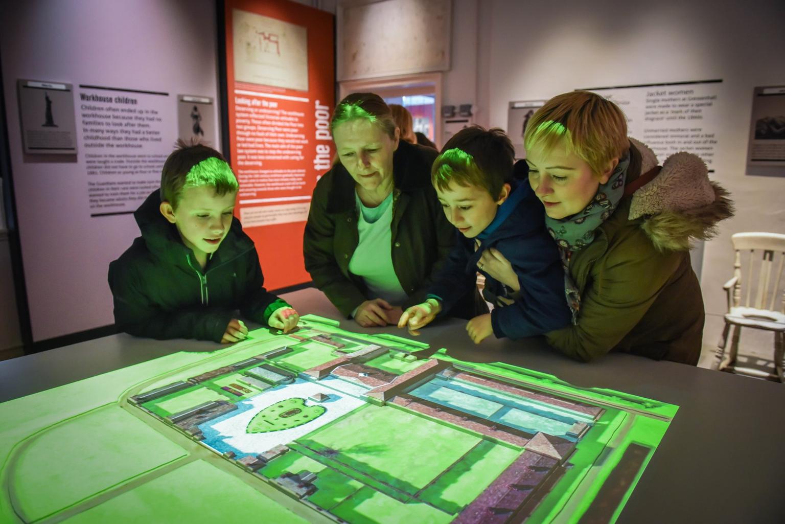 A family of two boys and two women lean over to look at a lit up aerial drawing of the farm's buildings and grounds.