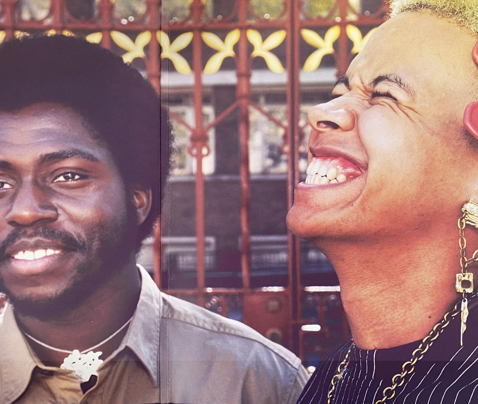 Two people stand in front of a red gate, one smiling towards the camera while the other with short bleached hair and a red afro comb grins with their head back.